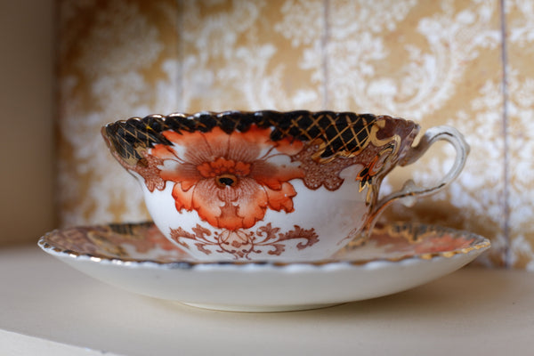 An ornate white porcelain teacup and saucer set with a large orange floral design, gold lattice detailing, and a black scalloped rim, displayed on a white surface against a patterned beige and white background.