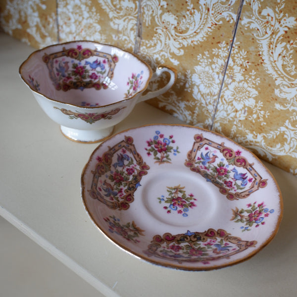 A vintage porcelain teacup and saucer set with a pink floral pattern and gold trim, displayed on a white surface against a gold damask wallpaper background.