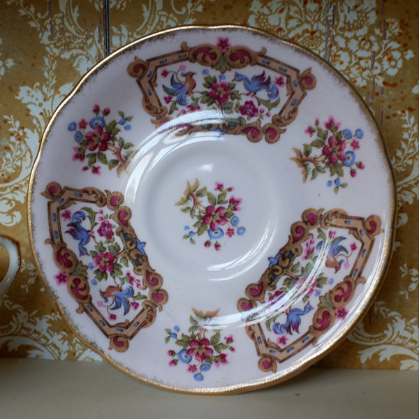 A vintage porcelain teacup saucer with ornate floral and scroll patterns in pink, blue, green, and gold, set against a gold damask wallpaper background.