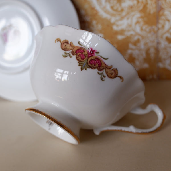 A white porcelain teacup with a floral scroll motif in pink, gold, and green, shown tipped on its side with its saucer behind it on a beige surface against a damask-patterned background.