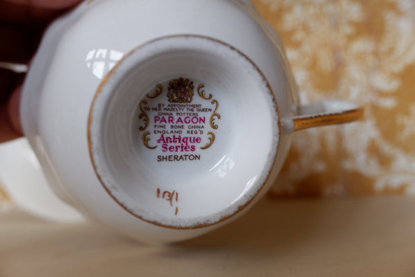 Close-up of the inside bottom of a white porcelain teacup or teapot base, showing printed text and crest for Paragon Antique Series Sheraton, with a gold rim and a hand holding it.