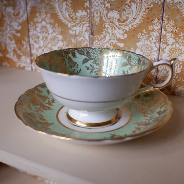 A white porcelain teacup and saucer set with a mint green damask pattern and gold trim, displayed on a shelf against a patterned wallpaper background.