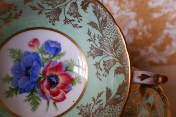 Close-up of an ornate porcelain teacup saucer with a mint green border, gold floral detailing, and a central floral medallion featuring blue and red anemone flowers.