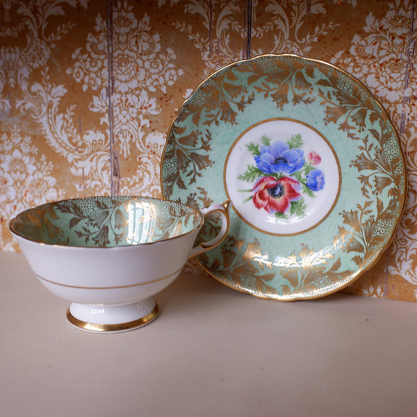 A vintage teacup and saucer set with a mint green and gold damask pattern, featuring a central floral motif with blue and red flowers, displayed against a gold and white damask wallpaper background.