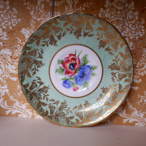 A vintage decorative ceramic plate with a gold and mint green floral border and a central floral motif of red and blue flowers, displayed against a patterned wallpaper background.