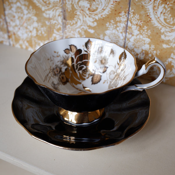A vintage teacup and saucer set with a glossy black exterior, gold trim, and a white interior decorated with gold floral patterns, set against a damask wallpaper background.