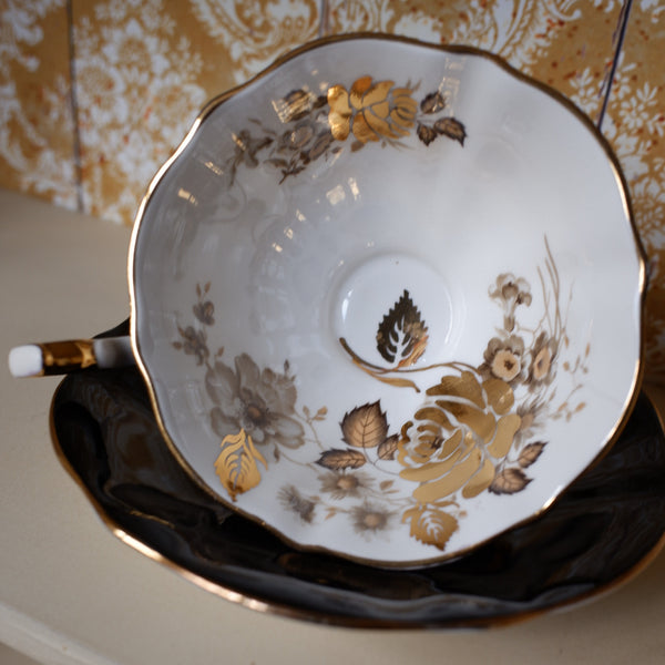 A white porcelain teacup and saucer set with gold and brown floral detailing, displayed on a beige surface against a patterned gold and white background.
