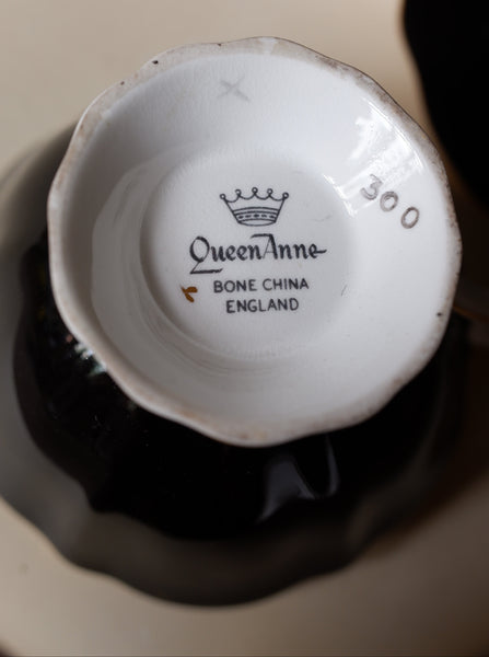 The bottom of a white ceramic teacup or small bowl with black printed branding reading 'Queen Anne BONE CHINA ENGLAND' and the number '300', shown resting on a dark saucer against a light beige background.