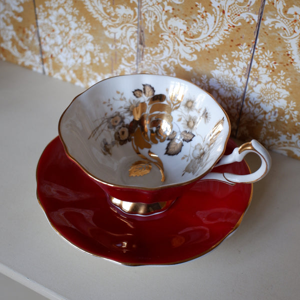 A vintage-style teacup and saucer set on a white surface, featuring a deep red saucer with gold trim and a white cup decorated with gold floral patterns and a gold handle.