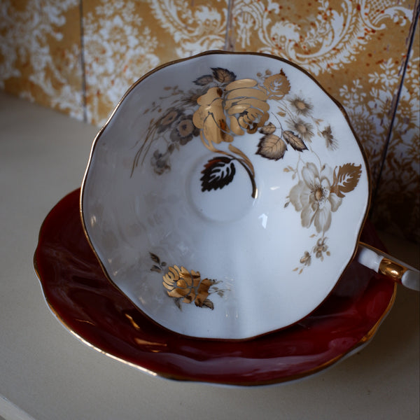 A white porcelain teacup with gold floral detailing, resting on a deep red saucer with gold trim, set against a patterned gold and white damask wallpaper background.