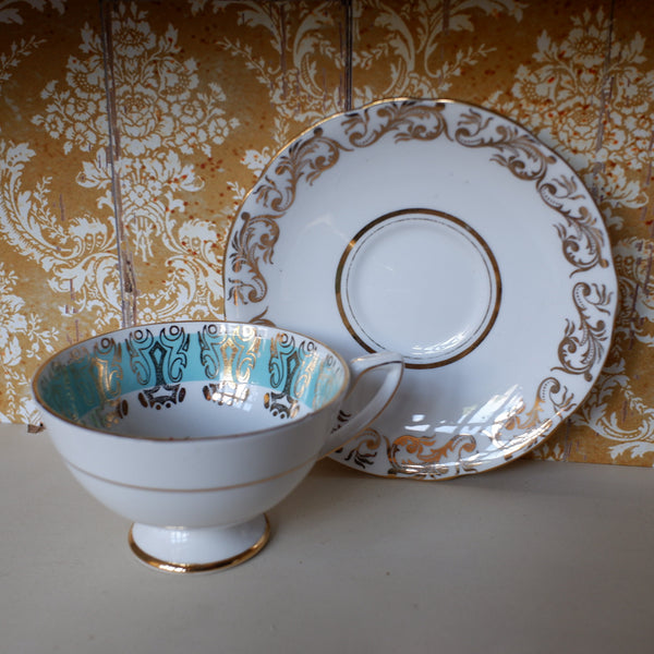 A white porcelain teacup and saucer set with ornate gold scrollwork and mint green patterned interior detailing, displayed against a patterned gold and white damask wallpaper background.
