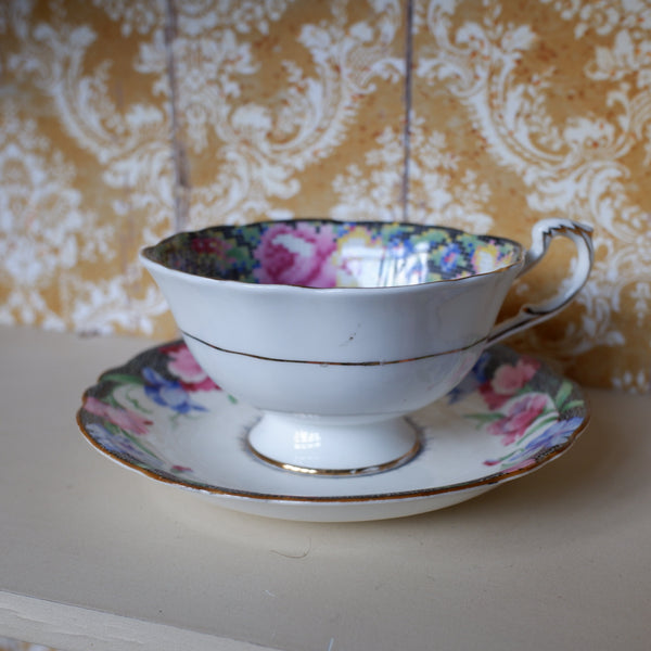 A white porcelain teacup and saucer set with a pink and blue floral pattern and gold trim, displayed on a light-colored wooden shelf against a damask wallpaper background.