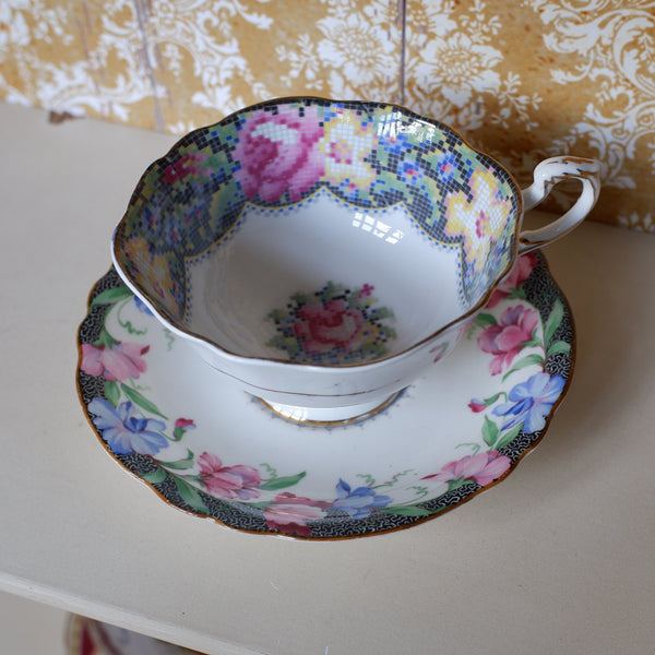 A vintage floral teacup and saucer set with pink roses, blue flowers, and gold trim, displayed on a light-colored shelf against a patterned wallpaper background.