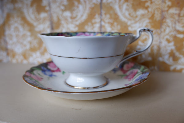 A vintage white porcelain teacup and saucer set with gold trim and a pink floral pattern, set against a patterned yellow and white damask background.