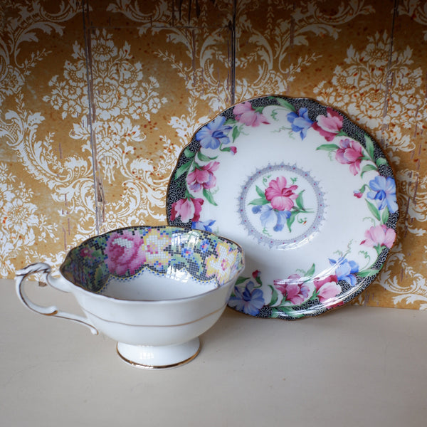 A vintage floral teacup and saucer set displayed on a shelf against a gold damask wallpaper background. The teacup is white with pink, blue, and green floral patterns, and the matching saucer features a central pink rose motif.