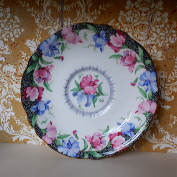 A round white porcelain plate with a colorful floral pattern, featuring pink and blue flowers with green leaves and a black speckled rim, set against a patterned gold and white wallpaper background.