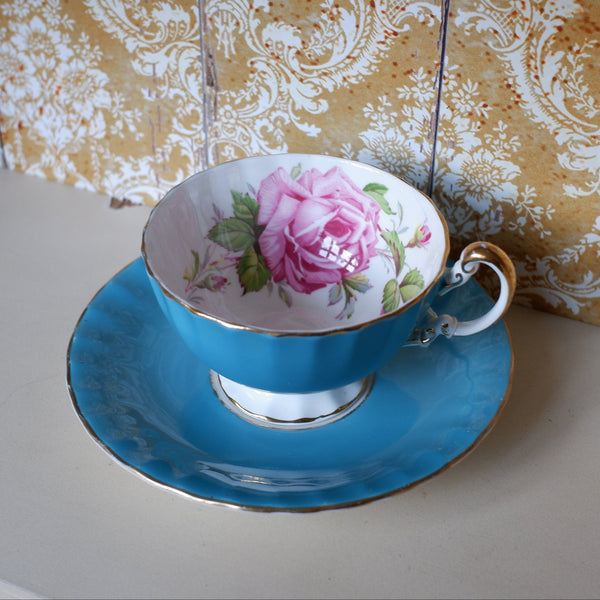 A vintage-style blue teacup and saucer set with a pink rose floral print on the inside of the cup, displayed on a shelf against a gold and white damask wallpaper background.