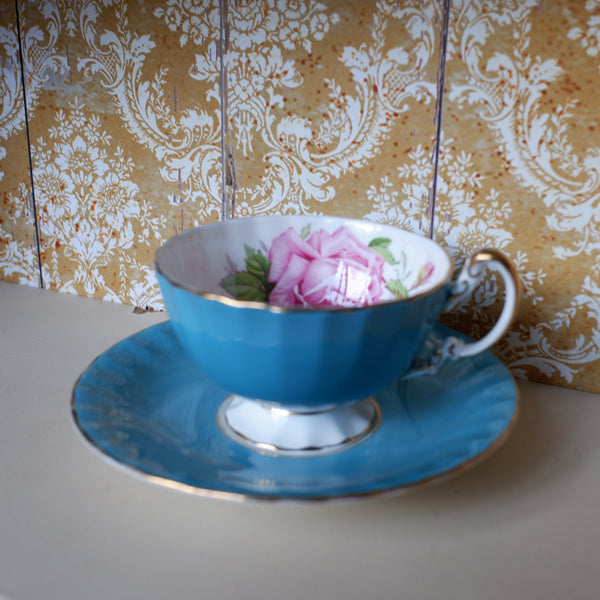 A vintage-style blue porcelain teacup and saucer set with gold trim, featuring a pink rose design on the inside, displayed on a light-colored surface against a patterned wallpaper background.