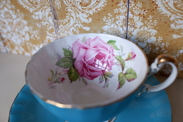 A vintage-style porcelain teacup with a pink rose and green leaves pattern, resting on a blue saucer against a patterned gold and white background.