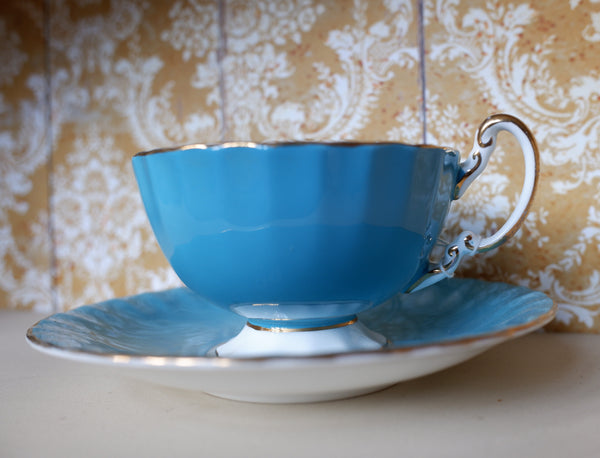A classic blue porcelain teacup and saucer set with gold trim, displayed on a light surface against a patterned gold and white damask wallpaper background.