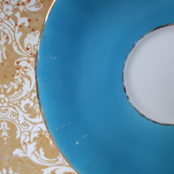 Close-up of a ceramic plate or saucer showing a bright turquoise blue interior, a white center, and part of a gold-and-white damask patterned rim.