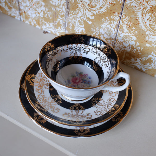An ornate white porcelain teacup and saucer set with black and gold damask patterns, featuring a small floral design on the inside of the cup, displayed on a light surface against a patterned gold and white background.