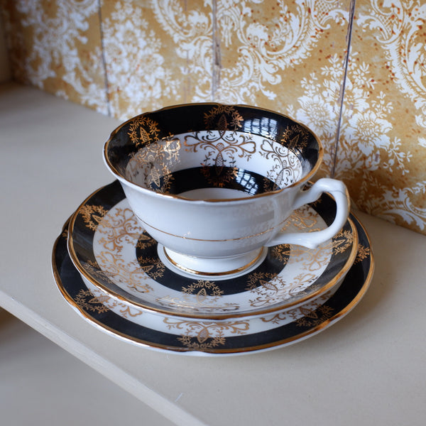 A white porcelain teacup and saucer set with ornate black and gold damask patterns, displayed on a light-colored shelf against a patterned gold and white wallpaper background.