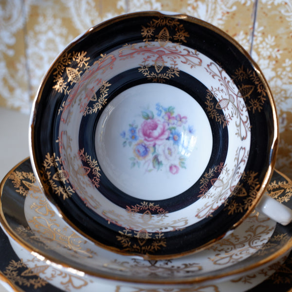 An ornate porcelain teacup and saucer set with a black and gold patterned rim, featuring a central floral design with pink, blue, and purple roses on a white background.