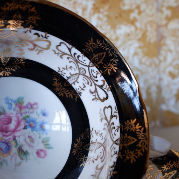 Close-up of an ornate white porcelain plate with a black and gold decorative border and a floral print in the lower left, set against a patterned gold and white background.