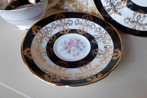 An ornate porcelain saucer with concentric black and white bands, gold damask-style detailing, and a central floral motif in pink, blue, and green, shown on a light tabletop with other matching dishes partially visible.