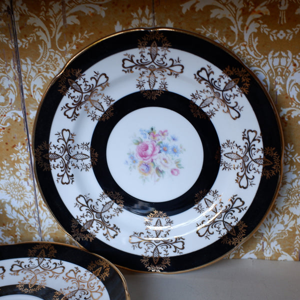 An ornate porcelain plate with a white center, black and gold scrollwork, and a pink floral motif, displayed against a gold damask wallpaper background.