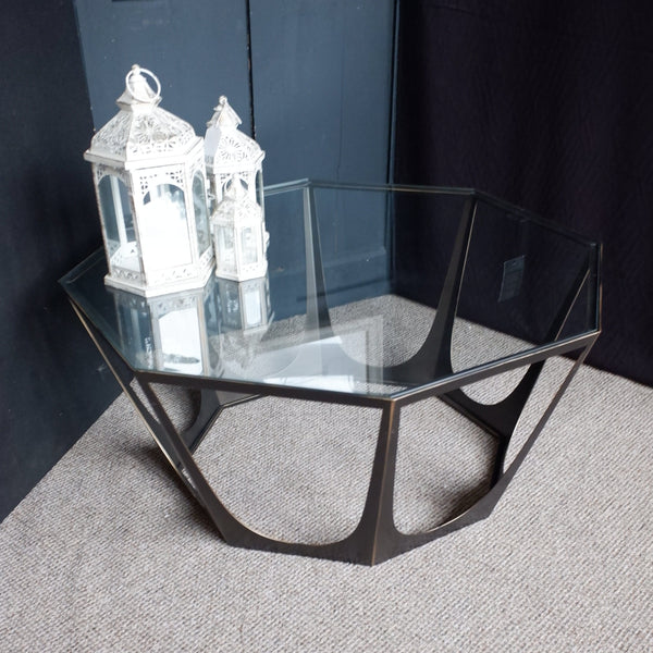 A modern octagonal coffee table with a clear glass top and a dark metal geometric frame, styled with two white lanterns on a textured gray carpet against a dark paneled wall.