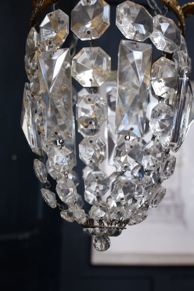 A close-up of a hanging crystal chandelier with faceted clear glass drops and clear crystal prisms, suspended from a gold-toned metal frame against a dark interior background.