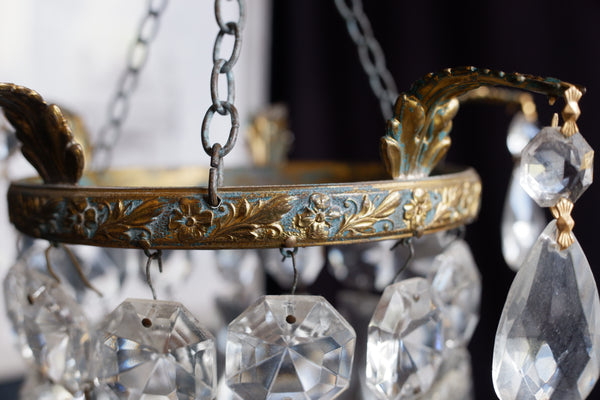 Close-up of an ornate hanging chandelier or lantern with a gold-toned floral metal frame, hanging chains, and clear faceted crystal drops.