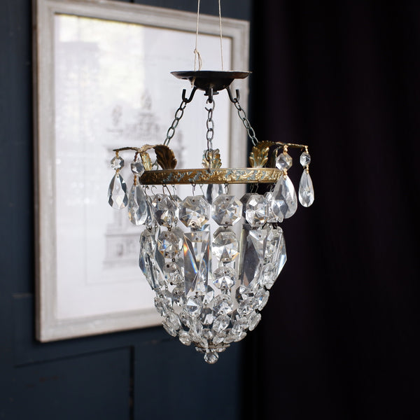 A vintage-style crystal chandelier pendant light with gold-toned metal detailing and clear glass teardrop prisms, hanging against a dark navy blue paneled wall next to a framed print.