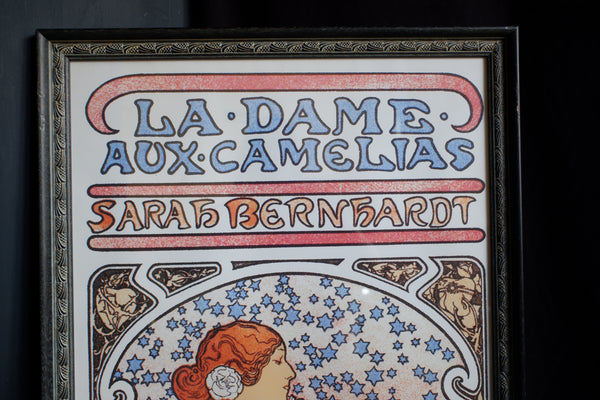 A framed vintage-style poster or print featuring the text 'LA DAME AUX CAMELIAS' and 'SARAH BERNHARDT' in decorative blue and orange lettering, with an Art Nouveau illustration below, shown against a dark background.