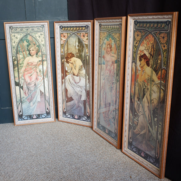 Four framed Art Nouveau style prints depicting female figures, arranged on a textured carpet against a dark paneled wall and black curtain.