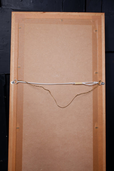 The back of a wooden framed art piece, showing a light brown cardboard backing secured with a metal wire and ring system against a dark background.
