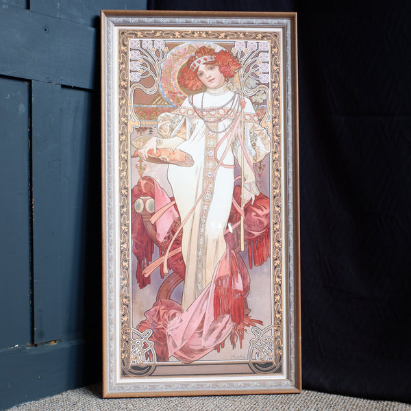 A framed Art Nouveau style print featuring an elegant woman in flowing white and pink garments, leaning against a dark blue wooden door and black fabric backdrop on a textured beige surface.