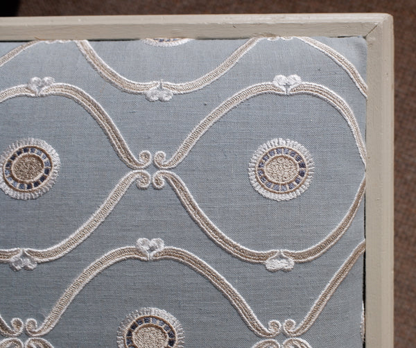 Close-up of a decorative fabric panel with a light blue background, white embroidered scrollwork, and circular medallion motifs, bordered by a cream-colored frame against a brown textured backdrop.