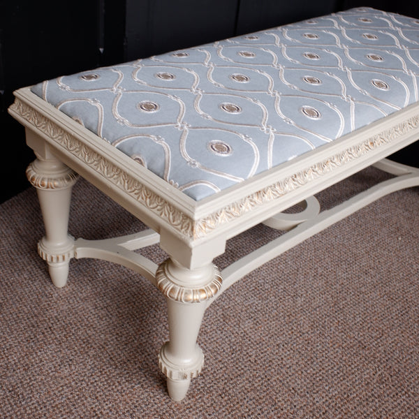 An ornate upholstered bench with a cream-colored carved frame and a light blue patterned cushion, set on a brown carpet against a dark wooden background.