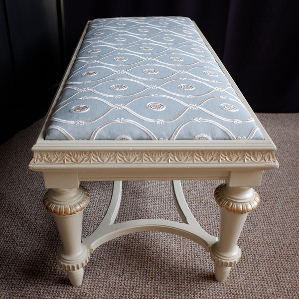 An ornate bench with a light blue patterned upholstered seat and a cream-colored carved wooden frame, set on a textured brown carpet against a dark background.