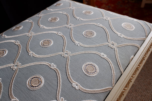 Close-up of a upholstered ottoman or bench cushion with a light blue fabric and ornate cream embroidery in a repeating geometric pattern, with part of the carved wooden frame visible on the right.