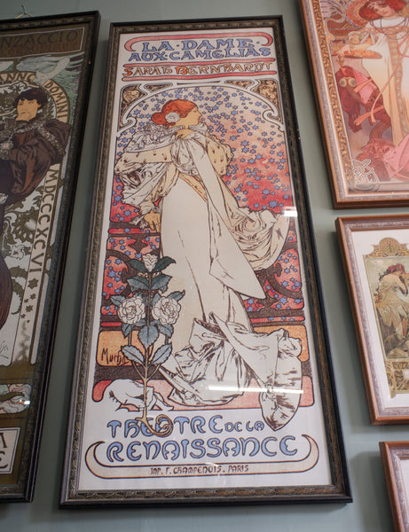 A framed Art Nouveau poster titled 'La Dame de Figarde' hangs on a gray wall, surrounded by other framed posters. The central poster features an elegant woman in flowing white and red dress with floral details and decorative typography.