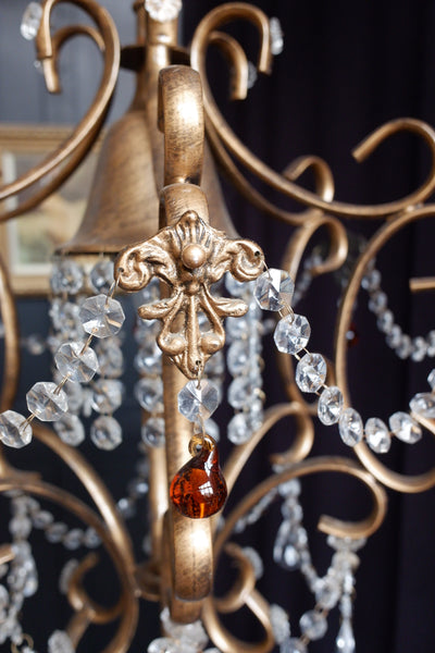 Close-up of an ornate gold-toned metal chandelier or candlelight fixture with clear hanging crystal beads and a single a single red teardrop pendant.