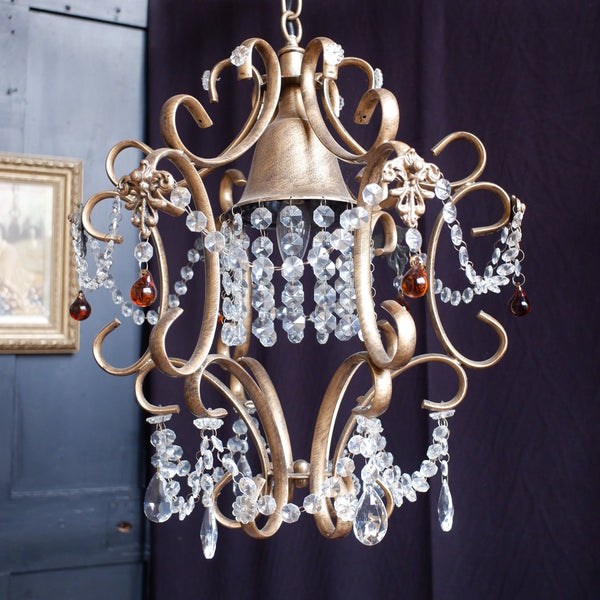 An ornate vintage-style brass chandelier with clear crystal droplets and red glass accents, hanging against a dark fabric backdrop.