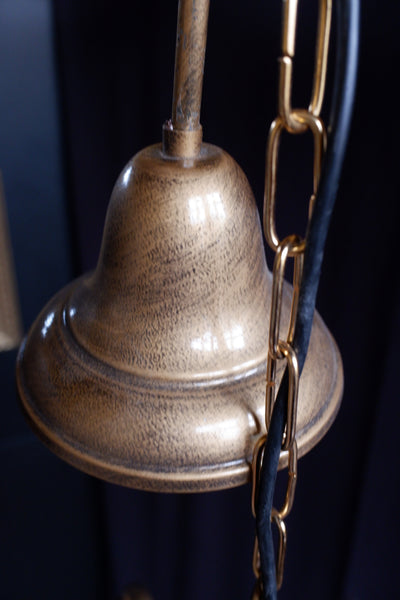 Close-up of a vintage-style bronze bell with a textured metallic finish, hanging from a chain with a black cord against a dark background.