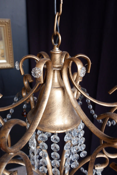 Close-up of an ornate chandelier with a distressed gold/bronze metal frame, curved scrollwork arms, and hanging clear faceted crystal beads.