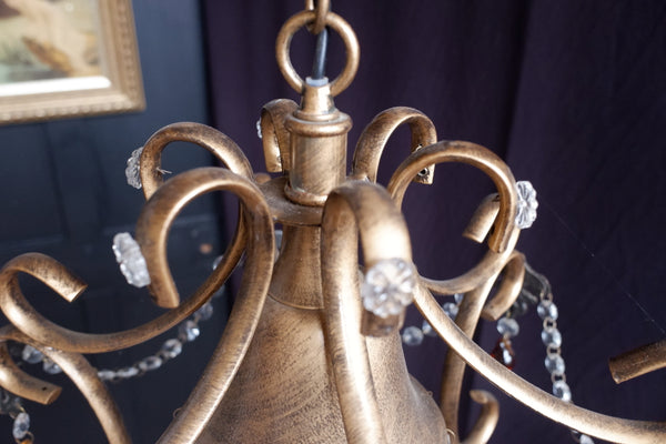 Close-up of an ornate chandelier fixture with curved metallic arms, a warm gold/brass finish, and hanging clear crystal bead strands against a dark purple fabric background.