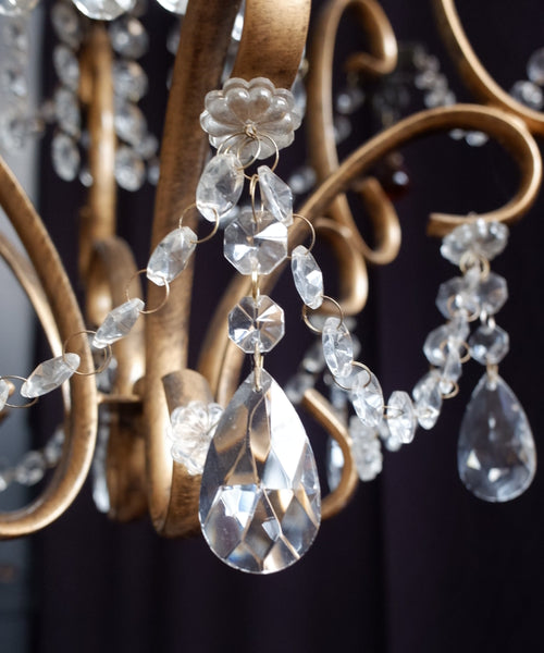 Close-up of a gold-toned chandelier with clear faceted crystal drops and teardrop pendants against a dark background.